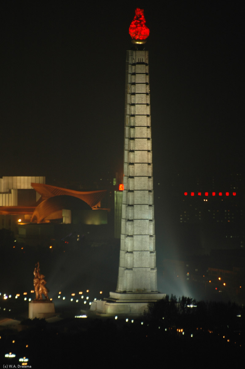 Another bright beacon in nightly Pyongyang is the lit Juche Tower. Even during power cuts the tower supposedly remains illuminated. In the back the roof of the East Pyongyang Grand Theatre is just visible.