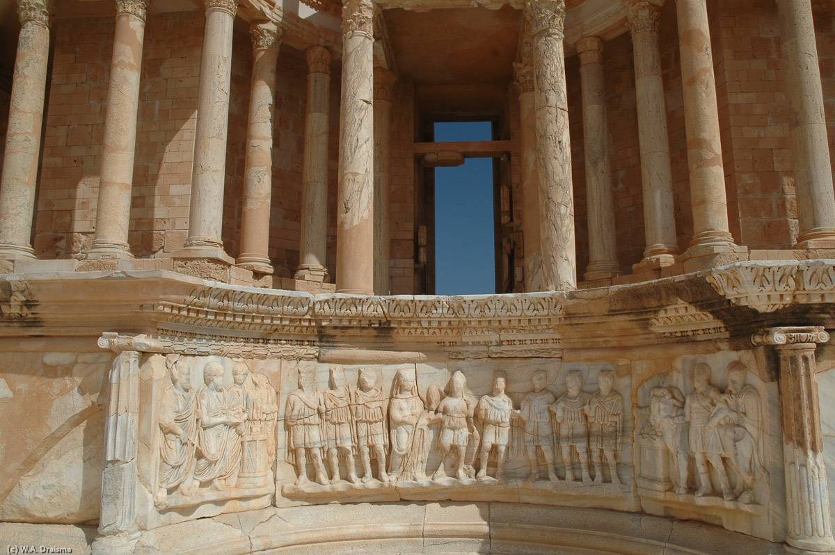 The elevated stage has three concave niches with marble panels. The central curved panel shows personifications of Rome and Sabratha, flanked by military figures and scenes of sacrifice.