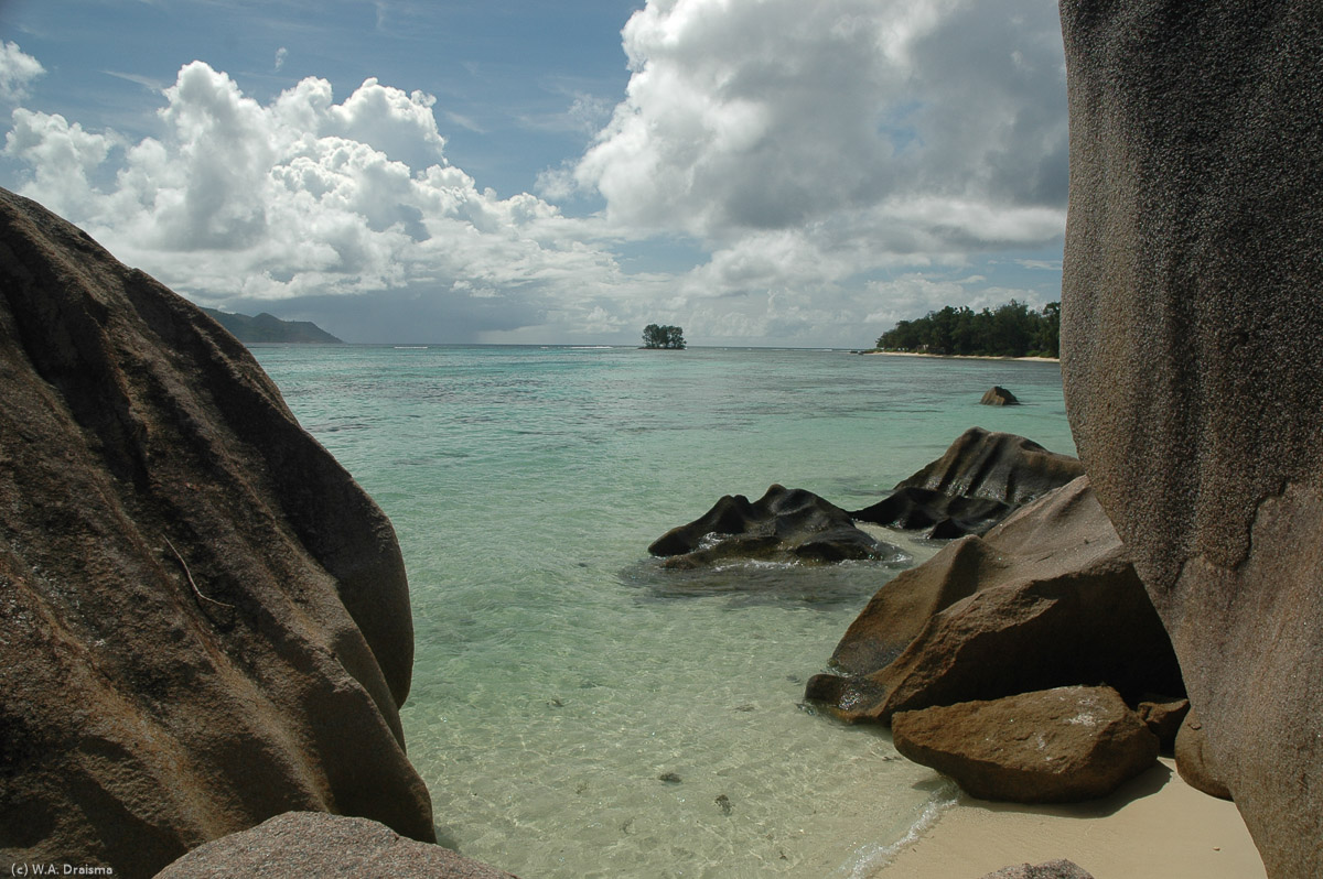 A peek between two huge boulders reveals a turqoise sea and, in the distance, La Passe (on the right) and Praslin (on the left).