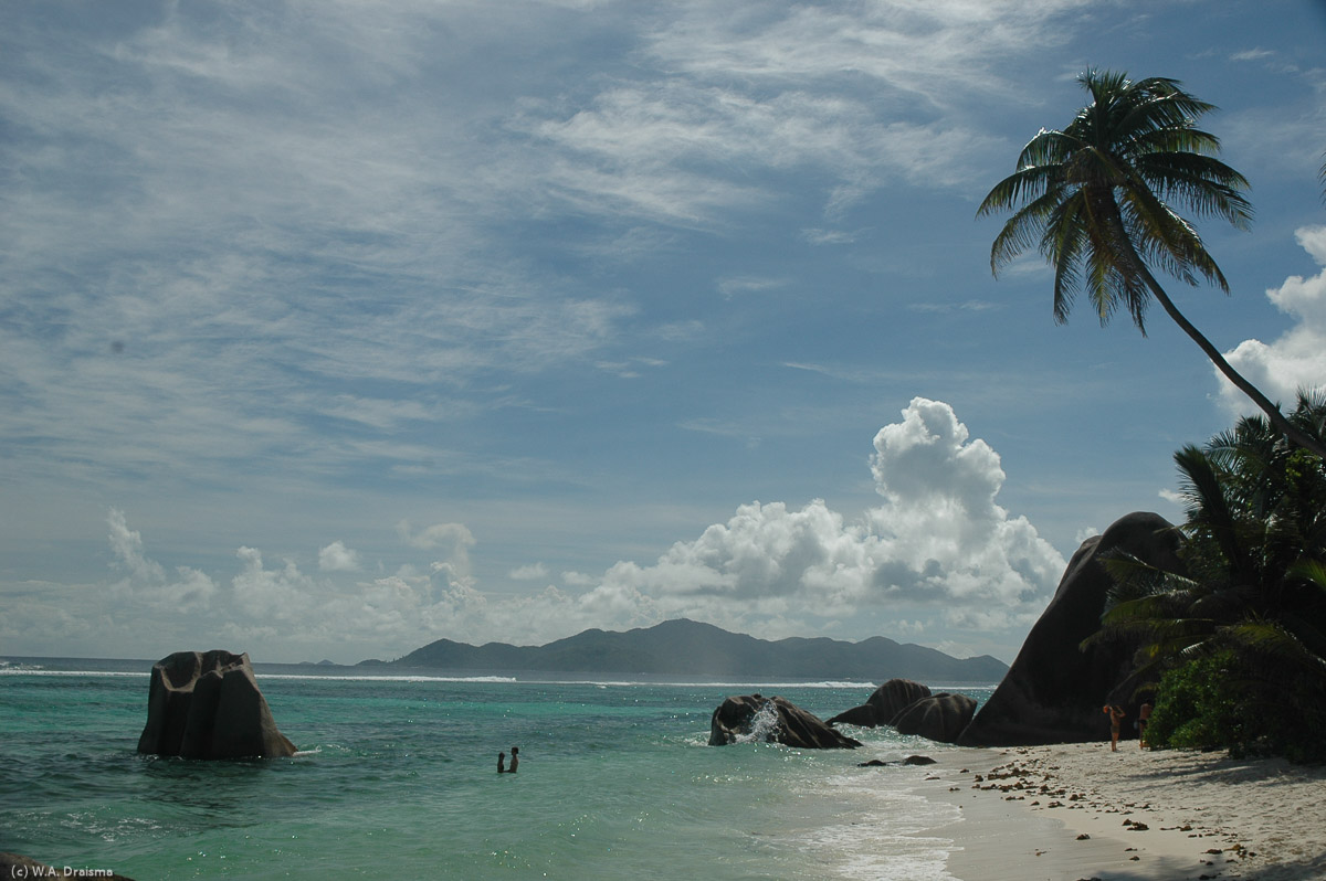 A couple enjoys the silence with only a coconut palm, granite boulders and Praslin in the distance as onlookers.